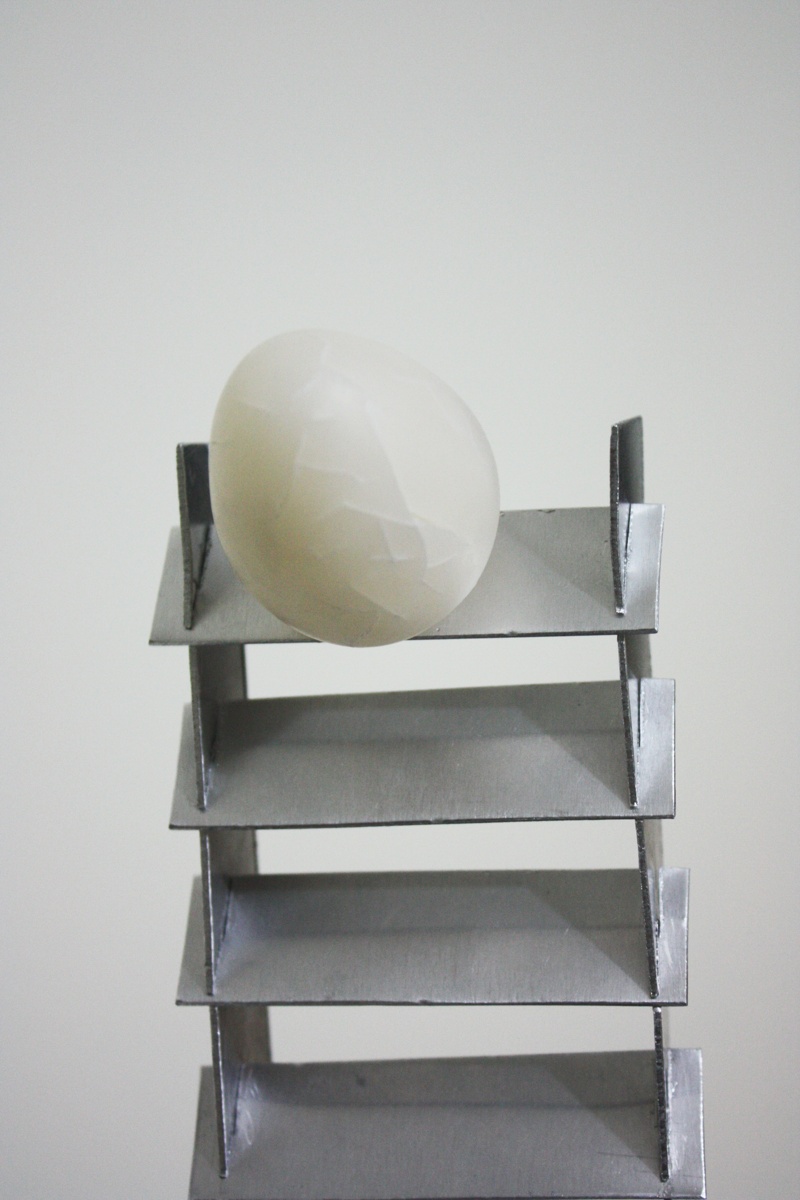 Close up of On the Verge sculpture by artist Priti Patel. It shows a fragile translucent cracked egg, made from cernit polymer clay, standing at the top of a twisted aluminium stair case that is tapered. The staircase is standing in a square pool of black oil. 